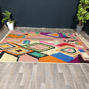 May include: A colorful geometric rug with a pink background and various shapes in orange, yellow, green, and blue. The rug has a fringed edge and is placed on a wooden floor.