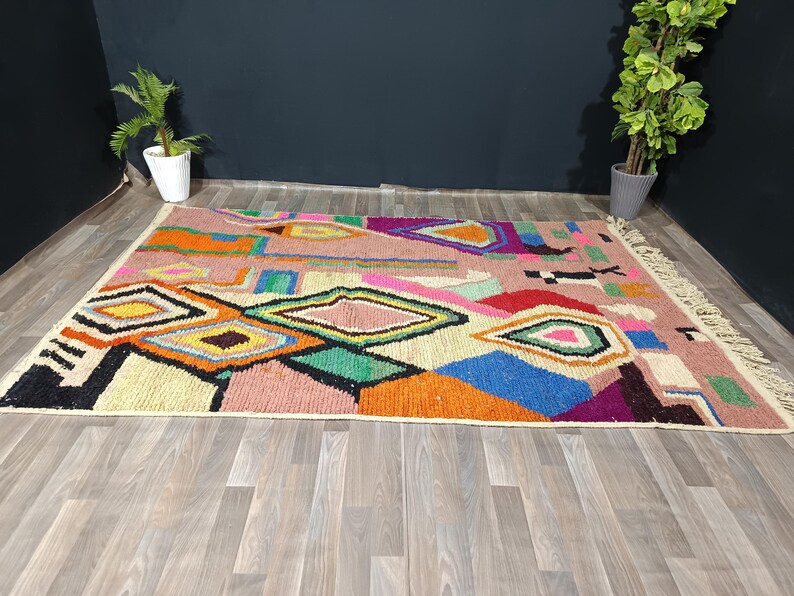 May include: A colorful geometric rug with a pink background and various shapes in orange, yellow, green, and blue. The rug has a fringed edge and is placed on a wooden floor.