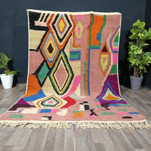 May include: A colorful, geometric rug with a pink background and a variety of shapes in orange, green, yellow, and blue. The rug has a fringe along the bottom edge and is displayed on a wooden floor.