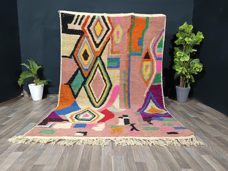 May include: A colorful, geometric rug with a pink background and a variety of shapes in orange, green, yellow, and blue. The rug has a fringe along the bottom edge and is displayed on a wooden floor.
