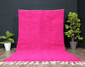 May include: A vibrant pink rug with a textured surface. The rug has a rectangular shape and is displayed on a wooden floor. White tassels adorn the bottom edge. The rug is a bold statement piece for any room.