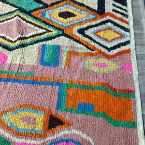May include: A colorful, geometric patterned rug with a mix of pink, orange, green, blue, and yellow diamond and rectangular shapes. The rug has a textured, woven appearance, with a cream-colored border.