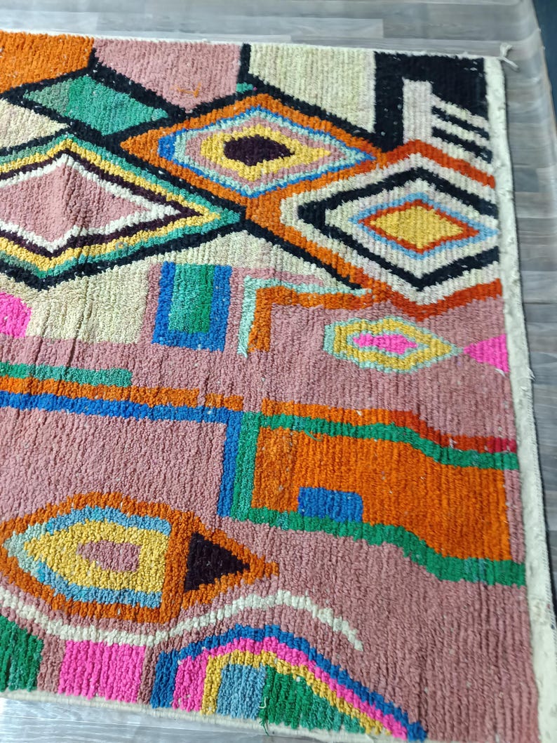 May include: A colorful, geometric patterned rug with a mix of pink, orange, green, blue, and yellow diamond and rectangular shapes. The rug has a textured, woven appearance, with a cream-colored border.