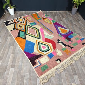 May include: A rectangular, hand-woven rug with a pink background and a geometric pattern in various colors. The design includes diamonds, squares, and other shapes in orange, blue, green, and white. The rug has a fringe on the edges.