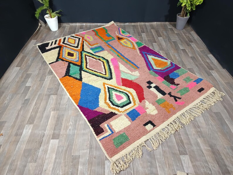 May include: A rectangular, hand-woven rug with a pink background and a geometric pattern in various colors. The design includes diamonds, squares, and other shapes in orange, blue, green, and white. The rug has a fringe on the edges.