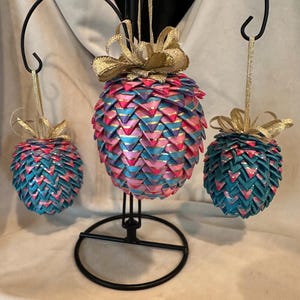 May include: Three decorative ornaments with a pineapple design. The ornaments are made of blue, pink, and gold materials. Each ornament has a gold ribbon bow at the top and is suspended by a gold string. The ornaments are displayed on a black metal stand.