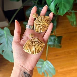 May include: A pair of gold-colored seashell-shaped earrings. The earrings have a textured, layered design, with a small round stud at the top. The earrings are displayed on a hand, with a silver ring visible.