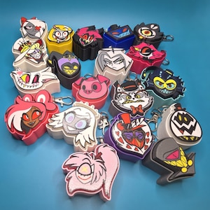 May include: A collection of multi-colored, three-dimensional character keychains featuring various animated faces with distinct expressions, colors, and shapes, each equipped with a metal lobster clasp for attachment.