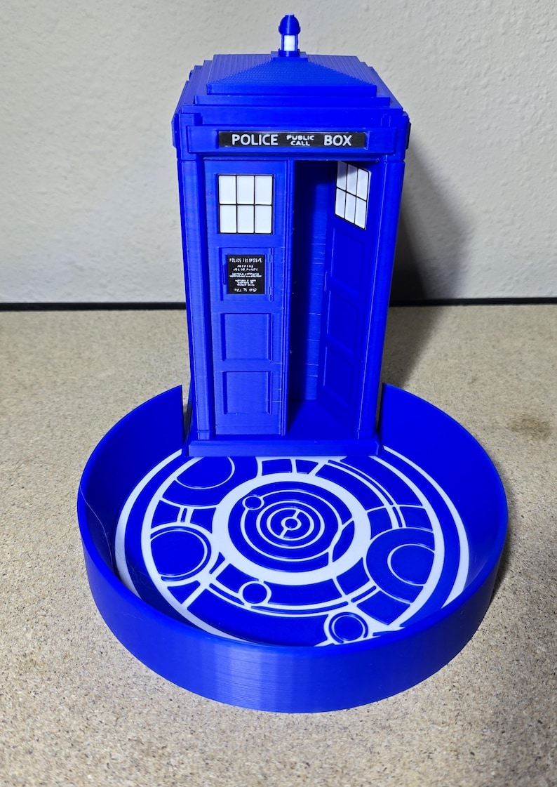 Puede incluir: Modelo azul impreso en 3D de una cabina de polic&iacute;a, con el texto "POLICE PUBLIC BOX" en la parte superior. Tiene una base circular con un dise&ntilde;o geom&eacute;trico blanco. La puerta est&aacute; abierta, revelando el interior.