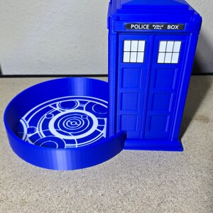Puede incluir: Un contenedor azul impreso en 3D con forma de Tardis con una bandeja circular. El contenedor tiene las palabras "POLICE PUBLIC BOX" encima de las puertas dobles. La bandeja presenta un dise&ntilde;o circular blanco.