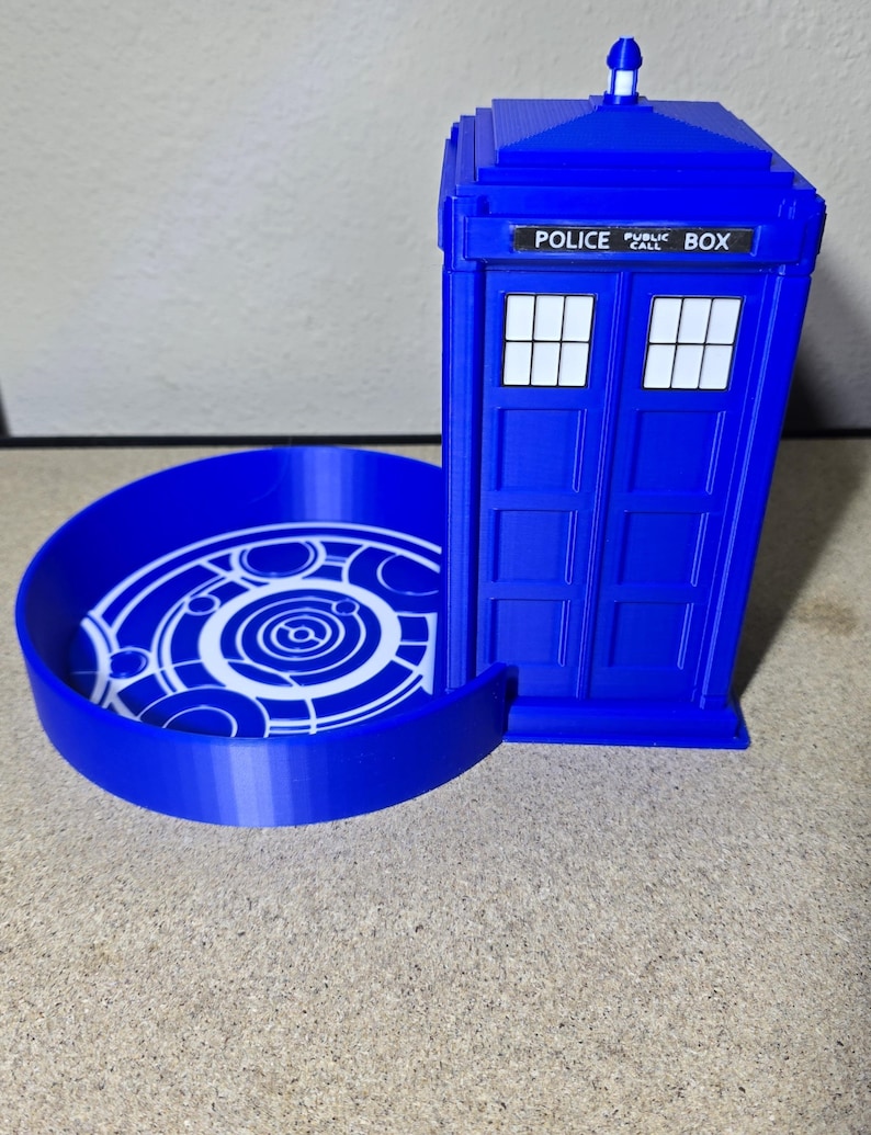 Puede incluir: Un contenedor azul impreso en 3D con forma de Tardis con una bandeja circular. El contenedor tiene las palabras "POLICE PUBLIC BOX" encima de las puertas dobles. La bandeja presenta un dise&ntilde;o circular blanco.