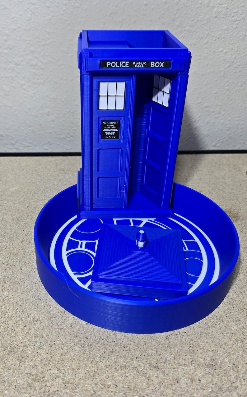 Puede incluir: Un modelo azul impreso en 3D de la ic&oacute;nica cabina de polic&iacute;a brit&aacute;nica, la TARDIS, de la serie Doctor Who. El modelo presenta el texto "POLICE PUBLIC CALL BOX" en la parte superior y est&aacute; sobre una base circular con detalles blancos.