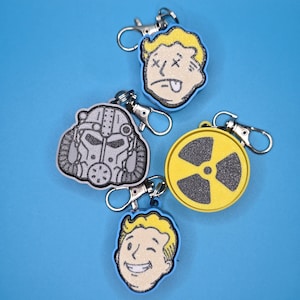 May include: Keychains featuring various designs, including a yellow and black radiation symbol, a gray helmet, and two different cartoon faces. Each keychain has a silver clasp and a blue border, set against a blue background.