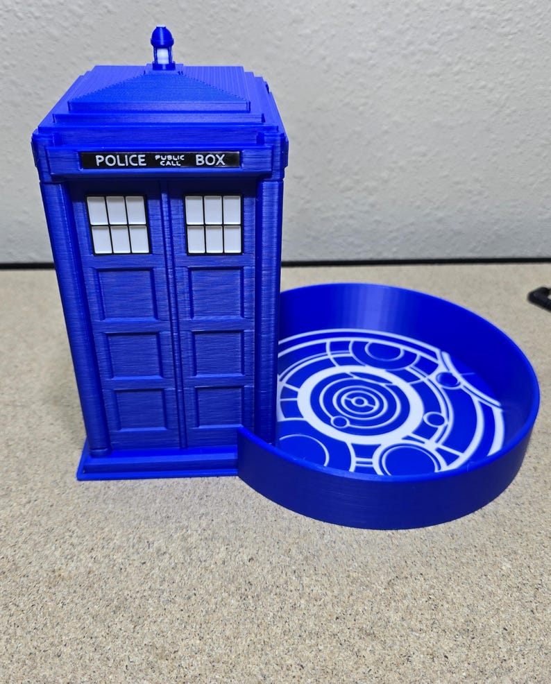 Puede incluir: Una cabina de polic&iacute;a azul impresa en 3D con una bandeja circular adjunta. La caja tiene las palabras "POLICE PUBLIC BOX" en la parte superior. La bandeja tiene un dise&ntilde;o circular blanco.