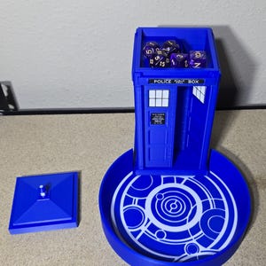 Puede incluir: Un juego de torre y bandeja para dados azules, que se asemeja a una cabina telef&oacute;nica de la polic&iacute;a. La torre tiene una puerta y ventanas, con las palabras "POLICE BOX" en la parte superior. La bandeja tiene un dise&ntilde;o circular. Dados morados est&aacute;n dentro.