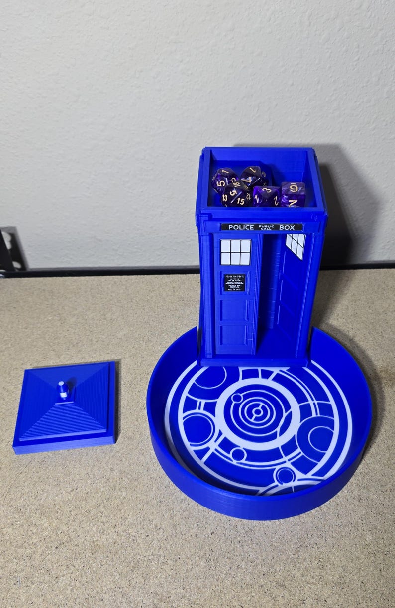 Puede incluir: Un juego de torre y bandeja para dados azules, que se asemeja a una cabina telef&oacute;nica de la polic&iacute;a. La torre tiene una puerta y ventanas, con las palabras "POLICE BOX" en la parte superior. La bandeja tiene un dise&ntilde;o circular. Dados morados est&aacute;n dentro.