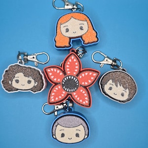 May include: A collection of character-themed keychains. The keychains feature cartoon-style illustrations of characters with glitter accents. The keychains are in various shapes, including a flower and character portraits, with silver-colored clasps.