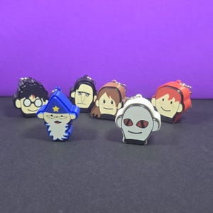 May include: A collection of six character-themed keychains featuring stylized heads. The figures include a boy with glasses, a blue wizard with a star, a man with dark hair, a person with brown hair, a white creature with red eyes, and a red-haired figure.