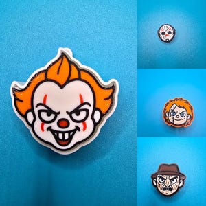 May include: A collection of four horror-themed character charms featuring a clown with orange hair, a white hockey mask, a doll with orange hair, and a figure in a brown hat. Each charm is rendered in a cartoon style with bold outlines and colors.