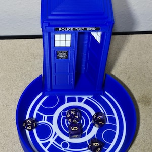 Puede incluir: Una bandeja para dados y una torre para dados azules, impresas en 3D, dise&ntilde;adas como una cabina telef&oacute;nica de la polic&iacute;a. La bandeja tiene un dise&ntilde;o circular blanco. Varios dados morados est&aacute;n dentro de la bandeja y la torre. La torre tiene las palabras "POLICE PUBLIC BOX".