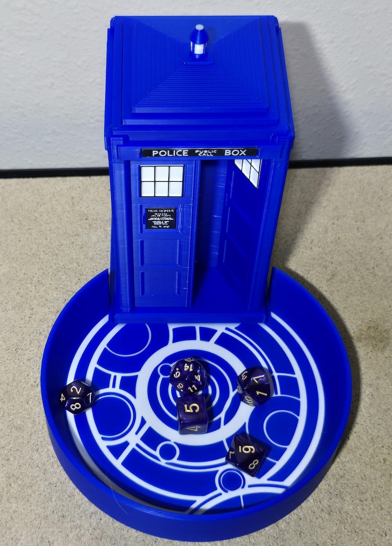 Puede incluir: Una bandeja para dados y una torre para dados azules, impresas en 3D, dise&ntilde;adas como una cabina telef&oacute;nica de la polic&iacute;a. La bandeja tiene un dise&ntilde;o circular blanco. Varios dados morados est&aacute;n dentro de la bandeja y la torre. La torre tiene las palabras "POLICE PUBLIC BOX".