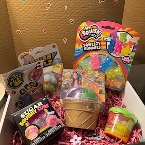 Puede incluir: Una caja de regalo llena de juguetes blandos y caramelos coloridos. La caja contiene una variedad de artículos, incluyendo gominolas, un cubo blando y un juguete de cono de helado de plástico. La caja está forrada con papel triturado rosa y decorada con pegatinas.
