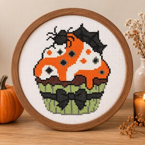 May include: A circular wooden embroidery hoop containing a cross-stitch pattern of a cupcake. The cupcake features orange and white frosting with black spider web accents, a dark brown base, and a light green wrapper with a black bow detail.