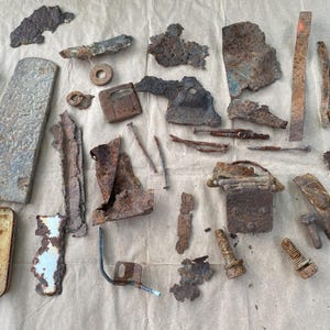 May include: A collection of assorted rusted metal fragments, including flat plates, bolts, nails, a coiled spring, and a small rectangular container. These weathered iron and steel pieces exhibit varying degrees of oxidation, textures, and irregular shapes.