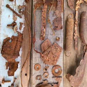 May include: A collection of assorted rusted metal hardware and scrap pieces, including bolts, washers, a coiled spring, and irregular flat metal fragments, all displaying heavy oxidation and a deep brown, textured, corroded surface finish.