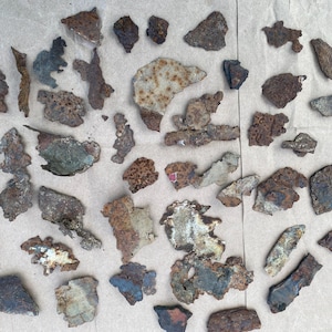 May include: A collection of numerous small, irregular metal fragments featuring oxidized surfaces with varying shades of brown, rust, gray, and metallic silver. Each piece displays a unique, jagged shape with a weathered, textured appearance.