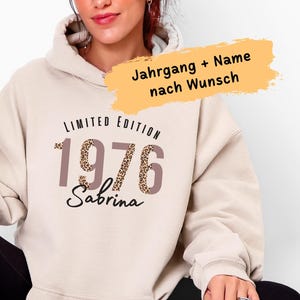 Könnte beinhalten: Beige Kapuzenpullover mit dem Text "LIMITED EDITION 1976 Sabrina" in Braun und Leopardenmuster. Die Person trägt schwarze Leggings, weiße Socken und Turnschuhe und hält einen Glasbecher mit Bambusdeckel.
