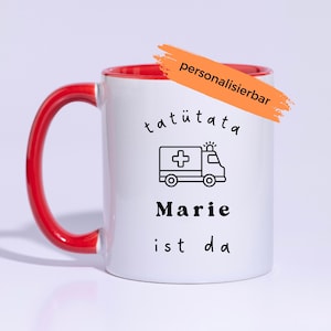 Könnte beinhalten: Eine weiße Keramiktasse mit rotem Innenraum und rotem Henkel. Auf der Vorderseite befindet sich eine schwarze Grafik eines Krankenwagens, der Text "tatütata", der Name "Marie" und der Text "ist da". Ein orangefarbener Pinselstrich mit dem Text "personalisierbar" ist zu sehen.