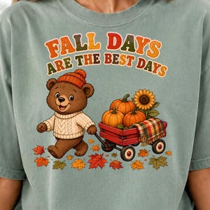 Può includere: Una t-shirt verde salvia con la grafica di un orso bruno che indossa un berretto arancione e un maglione color crema. L'orso tira un carro rosso pieno di zucche e un girasole. Il testo recita FALL DAYS ARE THE BEST DAYS sopra l'orso.