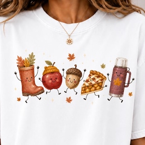 Può includere: Una t-shirt bianca a maniche corte con una stampa grafica di cinque personaggi animati a tema autunnale: uno stivale da pioggia con foglie, una mela rossa con cappello, una ghianda, una fetta di torta di zucca e un thermos marrone.