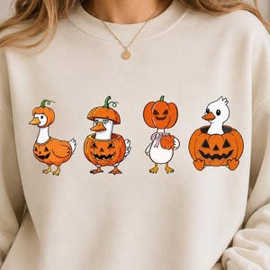 Può includere: Una felpa girocollo color crema con una stampa grafica di quattro oche bianche che interagiscono con zucche arancioni. Le oche sono raffigurate mentre indossano gusci di zucca come cappelli o sono sedute all'interno di zucche intagliate.