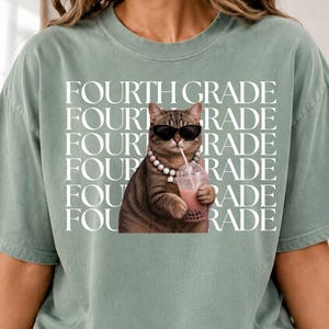 Può includere: Una t-shirt verde salvia con la grafica di un gatto soriano marrone che indossa occhiali da sole neri e una collana di perline mentre tiene in mano un bubble tea rosa. Il testo "FOURTH GRADE" è ripetuto in lettere maiuscole bianche.