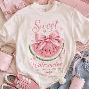 Può includere: T-shirt bianco con un disegno ad acquerello di una fetta di anguria e un fiocco rosa. Il testo "Sweet like a Watermelon" è scritto in rosa. La maglietta è abbinata a scarpe da ginnastica rosa e jeans.