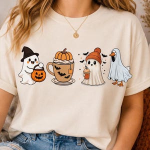 Può includere: Una t-shirt color crema con una stampa grafica di quattro personaggi a tema Halloween: un fantasma con cappello da strega e secchiello a forma di zucca, una tazza di tè alla zucca con pipistrelli, un fantasma con berretto arancione che tiene una bevanda e un'anatra in costume da fantasma.