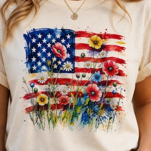 Può includere: T-shirt color crema con un disegno ad acquerello della bandiera americana, con strisce rosse e bianche, un campo di stelle blu e fiori colorati. I fiori includono papaveri rossi, margherite gialle e fiori blu. La maglietta ha maniche corte.