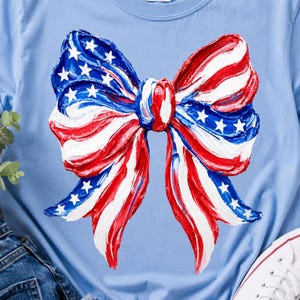 Può includere: T-shirt azzurro con un grande disegno a fiocco. Il fiocco è dipinto con strisce rosse e bianche e sezioni blu con stelle bianche, che ricordano la bandiera americana. La maglietta è realizzata in materiale morbido.