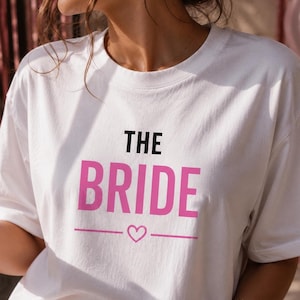 Può includere: Una t-shirt bianca a maniche corte con il testo nero THE sopra il testo rosa BRIDE, seguito da una linea orizzontale con un piccolo cuore rosa al centro e un'altra linea orizzontale, il tutto stampato sulla parte anteriore del capo.