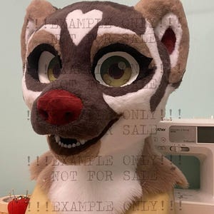 May include: A plush animal head mask featuring brown, tan, and white faux fur. The mask has large green eyes with black outlines, a dark red nose, and a white heart-shaped marking on the forehead. The mouth is slightly open, revealing white teeth.
