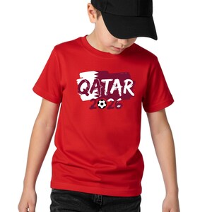 Puede incluir: Una camiseta roja con la palabra "QATAR" en blanco, sobre un dise&ntilde;o granate y blanco. El n&uacute;mero "2026" y un gr&aacute;fico de bal&oacute;n de f&uacute;tbol est&aacute;n debajo de la palabra "QATAR". La camiseta la lleva una persona con una gorra de b&eacute;isbol negra.