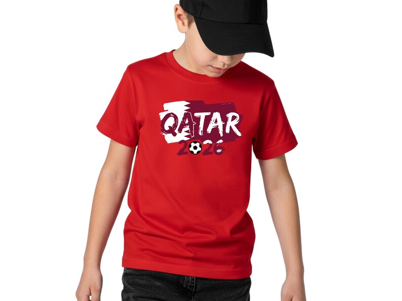 Puede incluir: Una camiseta roja con la palabra "QATAR" en blanco, sobre un dise&ntilde;o granate y blanco. El n&uacute;mero "2026" y un gr&aacute;fico de bal&oacute;n de f&uacute;tbol est&aacute;n debajo de la palabra "QATAR". La camiseta la lleva una persona con una gorra de b&eacute;isbol negra.
