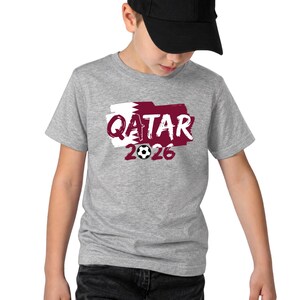 Puede incluir: Una camiseta gris con la palabra "QATAR" en borgo&ntilde;a, sobre un fondo blanco y borgo&ntilde;a, y el n&uacute;mero "2026" con un bal&oacute;n de f&uacute;tbol. La camiseta la lleva una persona con una gorra de b&eacute;isbol negra.