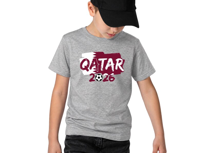 Puede incluir: Una camiseta gris con la palabra "QATAR" en borgo&ntilde;a, sobre un fondo blanco y borgo&ntilde;a, y el n&uacute;mero "2026" con un bal&oacute;n de f&uacute;tbol. La camiseta la lleva una persona con una gorra de b&eacute;isbol negra.