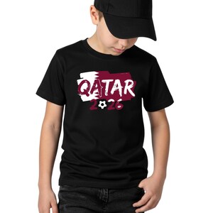 Puede incluir: Camiseta negra con la palabra "QATAR" en blanco, sobre un dise&ntilde;o granate y blanco, y el n&uacute;mero "2026" con un gr&aacute;fico de bal&oacute;n de f&uacute;tbol. La camiseta la lleva una persona con una gorra negra.