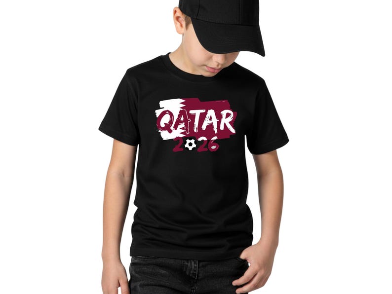 Puede incluir: Camiseta negra con la palabra "QATAR" en blanco, sobre un dise&ntilde;o granate y blanco, y el n&uacute;mero "2026" con un gr&aacute;fico de bal&oacute;n de f&uacute;tbol. La camiseta la lleva una persona con una gorra negra.