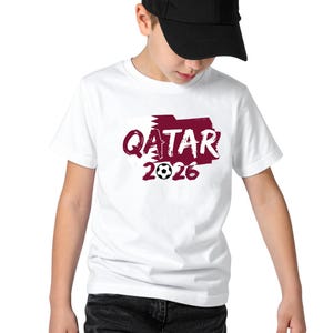 Puede incluir: Camiseta blanca con un gr&aacute;fico granate que dice "QATAR 2026" con un bal&oacute;n de f&uacute;tbol. La persona lleva una gorra de b&eacute;isbol negra.