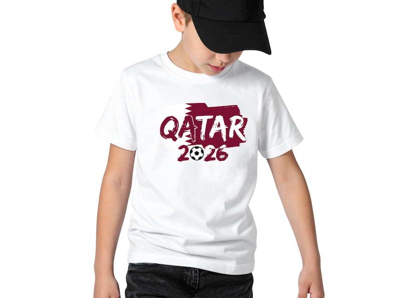 Puede incluir: Camiseta blanca con un gr&aacute;fico granate que dice "QATAR 2026" con un bal&oacute;n de f&uacute;tbol. La persona lleva una gorra de b&eacute;isbol negra.